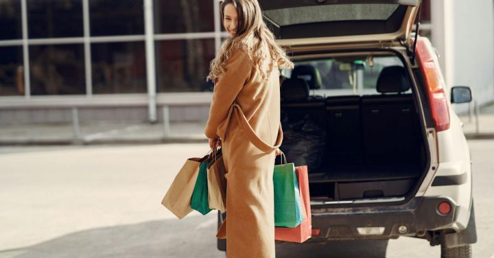 Sustainable Shopping - Full length of cheerful female looking over shoulder while standing near opened trunk of car on parking of shopping mall